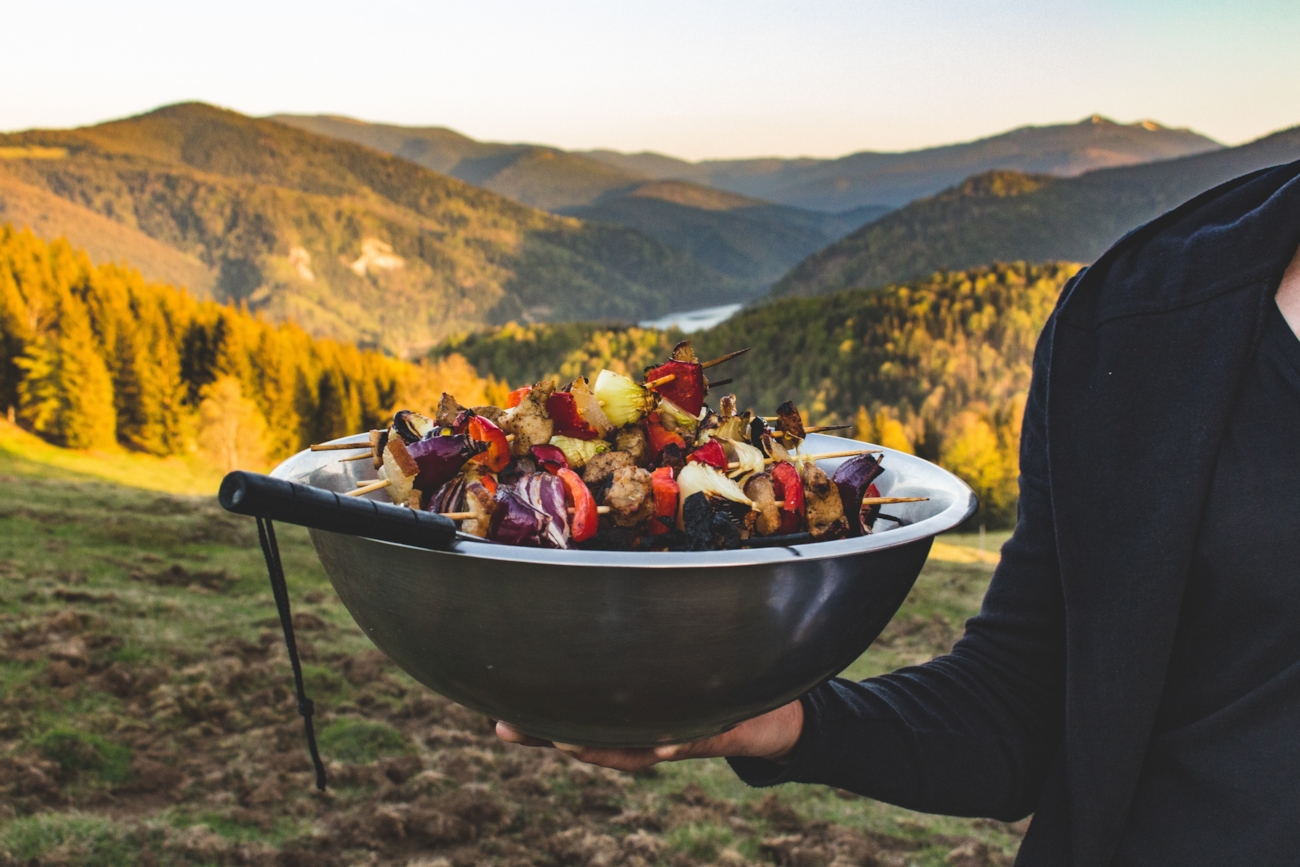 Hånd som holder en bolle med grillspyd på en bakketopp med skog i bakgrunnen